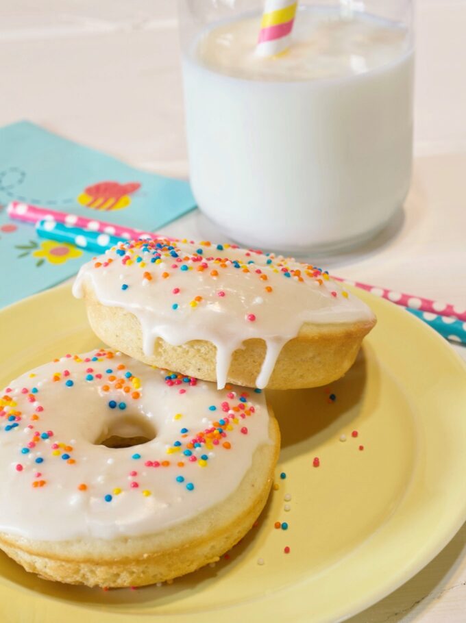 Two baked vanilla cake donuts with vanilla glaze and sprinkles on a yellow plate.